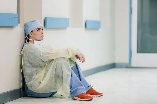 A tired doctor sits on the floor against a hospital wall